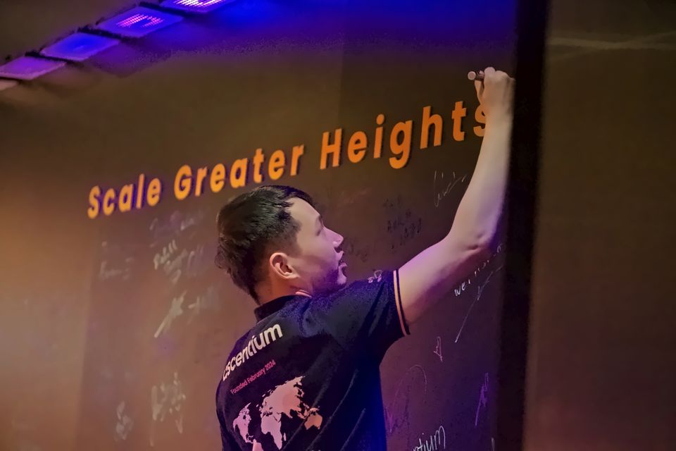 A guest in an Ascentium shirt signs the wall under the words Scale Greater Heights
