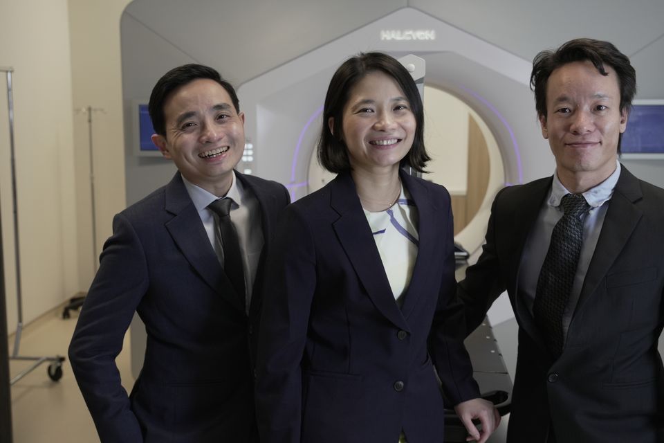 Three doctors in suits stand in front of the proton therapy machine