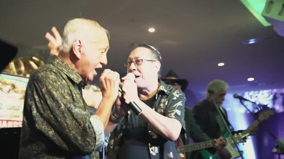 Two men sing together into one microphone in front of the band