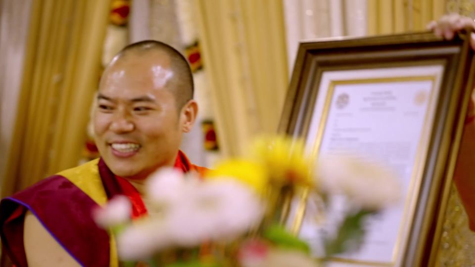 A smiling Rinpoche holds a framed certificate behind a vase of flowers